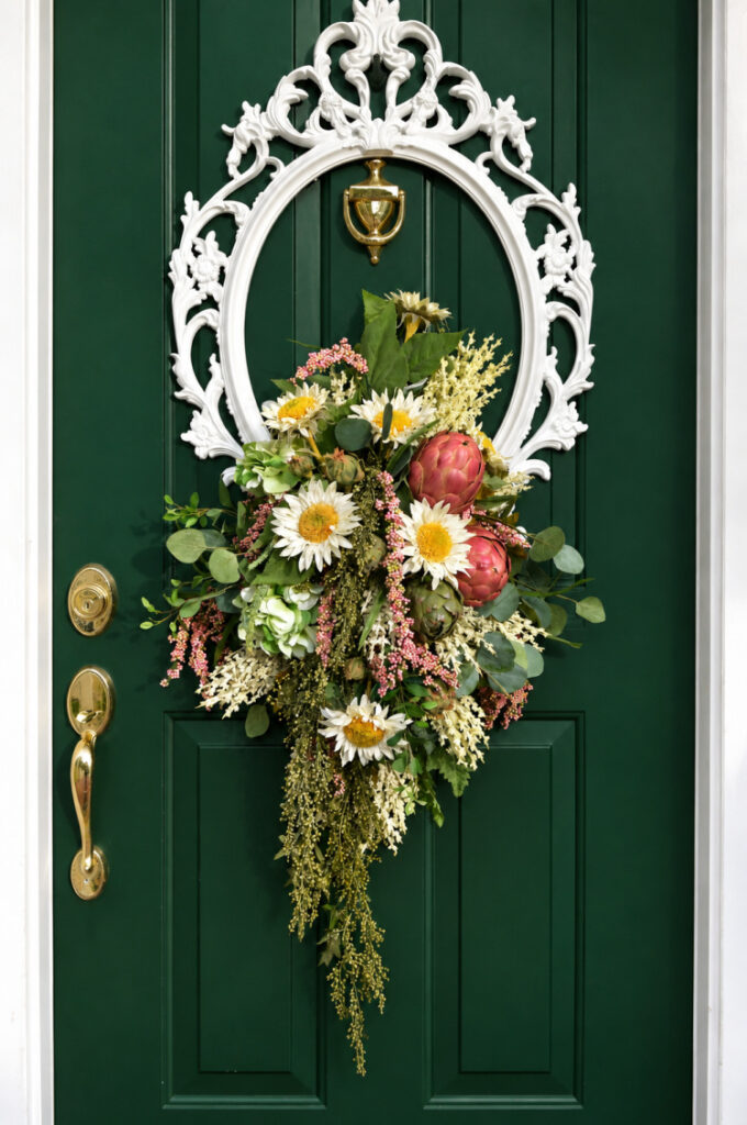 Finished DIY picture frame wreath with faux sunflowers, hydrangea, artichokes, eucalyptus, and cascading greenery arranged inside an ornate white frame on a green front door."