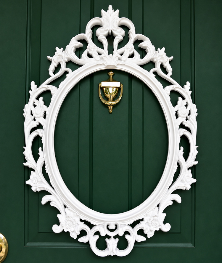 "Ornate white picture frame mounted on a dark green front door, creating the base for a DIY picture frame wreath."