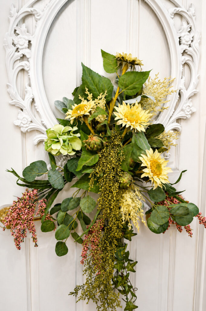 Finished DIY picture frame wreath with faux sunflowers, hydrangea, artichokes, eucalyptus, and cascading greenery arranged inside an ornate white frame on a white front door."