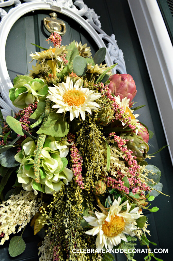 Finished DIY picture frame wreath with faux sunflowers, hydrangea, artichokes, eucalyptus, and cascading greenery arranged inside an ornate white frame on a green front door."