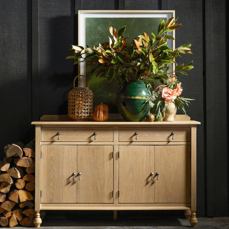 Light wood sideboard styled with a large green vase of magnolia branches, woven lantern, framed artwork, and seasonal accents for elegant late summer decorating.