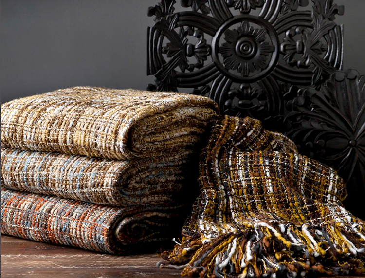 Stack of textured woven throw blankets in warm neutral and earthy tones displayed beside an ornate black carved chair, perfect for late summer and fall decorating.