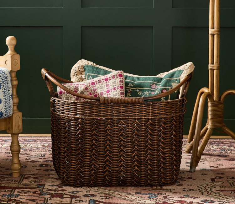 Dark brown woven wicker storage basket with curved wooden handles, styled with decorative pillows on a vintage-inspired rug against a deep green paneled wall.