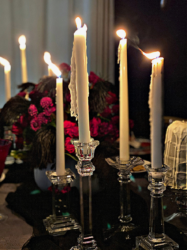 Dripping white taper candles in clear glass holders cast an eerie glow over a dark Halloween dinner-party table with deep red flowers and black feathers.