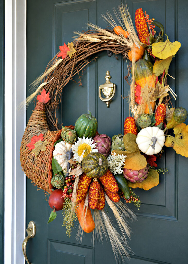Cornucopia-style fall wreath overflowing with pumpkins, gourds, artichokes, berries, autumn leaves, and wheat on a dark green front door.