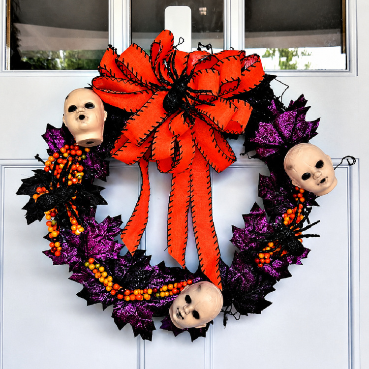 Halloween wreath with creepy doll heads, black spiders, glittered purple leaves, orange berries, and a large orange-and-black bow.