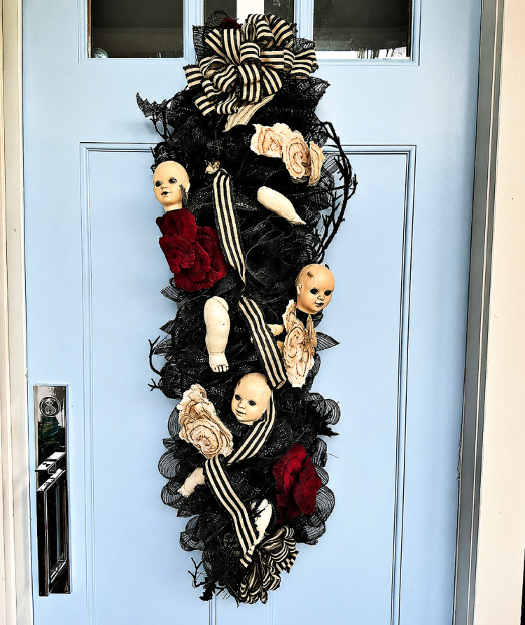 Halloween door swag featuring black mesh, striped ribbon, burgundy and cream flowers, and vintage doll heads and limbs displayed on a pale blue front door.