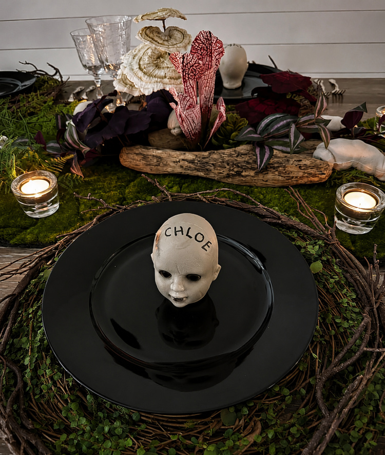 Spooky Halloween place setting with a creepy doll-head place card labeled “Chloe,” black dinnerware, moss, twisted branches, candlelight, and dramatic dark florals.