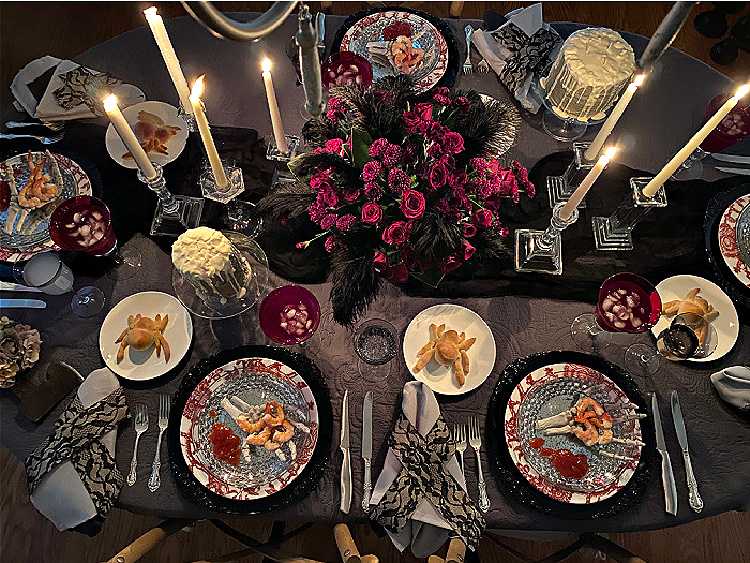 Overhead view of an elegant Halloween dinner table with glowing taper candles, patterned place settings, skeletal hands, and a dramatic centerpiece of red flowers and black feathers.