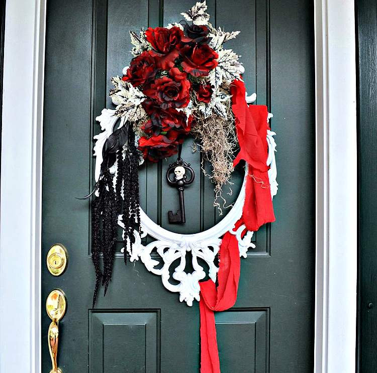 Ornate white picture-frame Halloween wreath decorated with red and black roses, pale foliage, Spanish moss, trailing black flowers, and tattered red fabric on a dark front door.