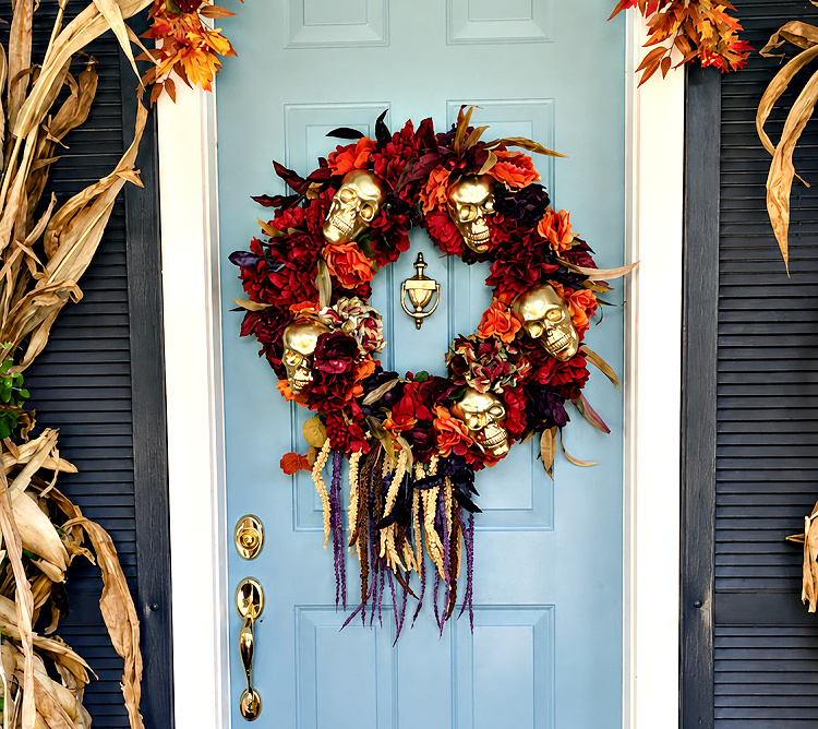 Lush Halloween wreath with orange and deep red flowers, metallic gold skulls, dark foliage, and trailing stems displayed on a pale blue front door.