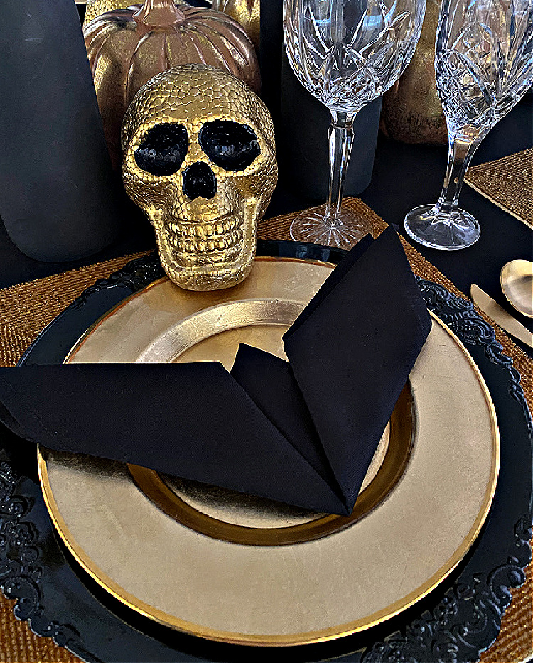 Elegant black-and-gold Halloween place setting with layered dinnerware, a folded black napkin, crystal glasses, metallic pumpkins, and a gold skull.