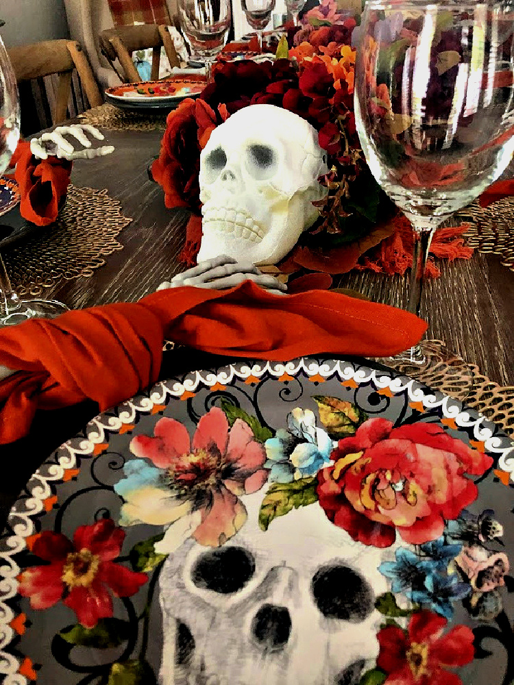 Halloween tablescape with a floral skull dinner plate, orange napkin, skeleton hands, and a flower-filled table runner accented with white skulls and bones.