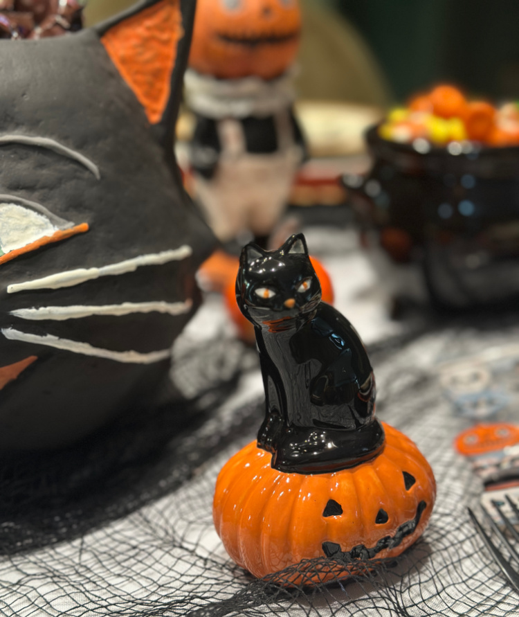 Vintage-style black cat figurine perched on an orange jack-o’-lantern beside a large black cat decoration on a Halloween table.