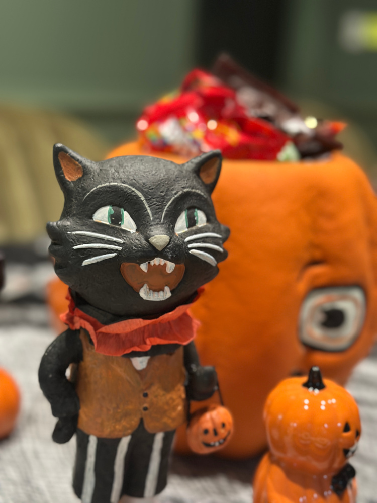 Vintage-style black cat figurine wearing a vest and holding a tiny jack-o’-lantern pail on a Halloween candy table.