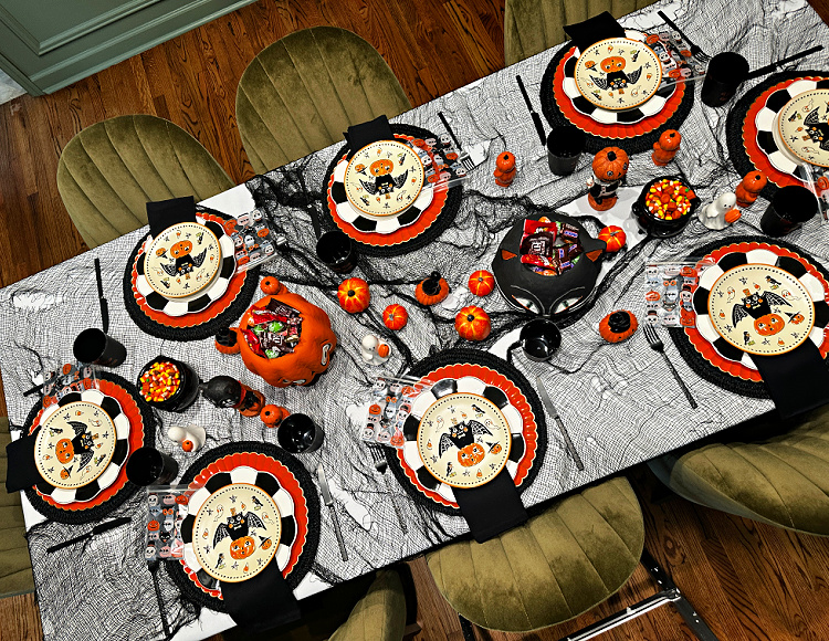 Overhead view of a vintage Halloween table set for eight with layered black-and-orange dishes, picture treat bags, candy bowls, pumpkins, and black gauze.