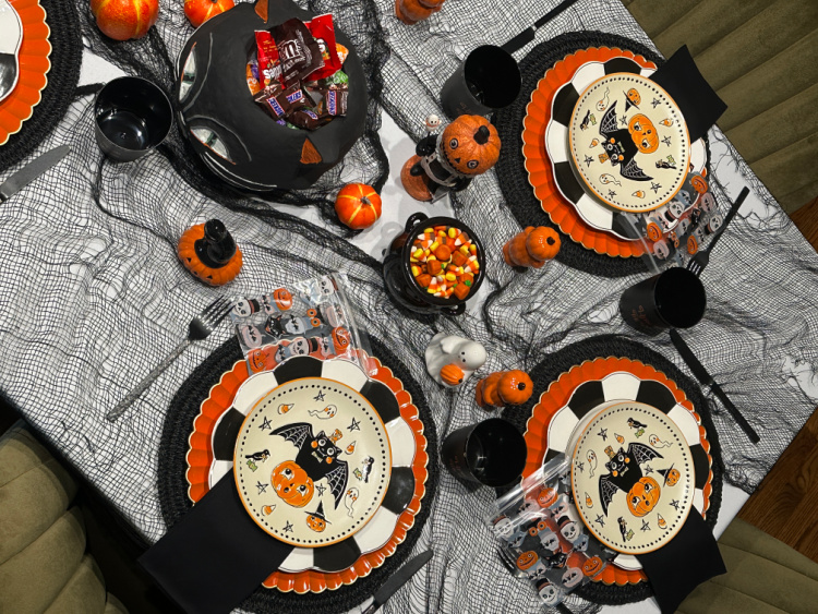 Overhead view of a vintage Halloween table with layered black, orange, and checkered dishes, picture treat bags, candy, pumpkins, and playful figurines.