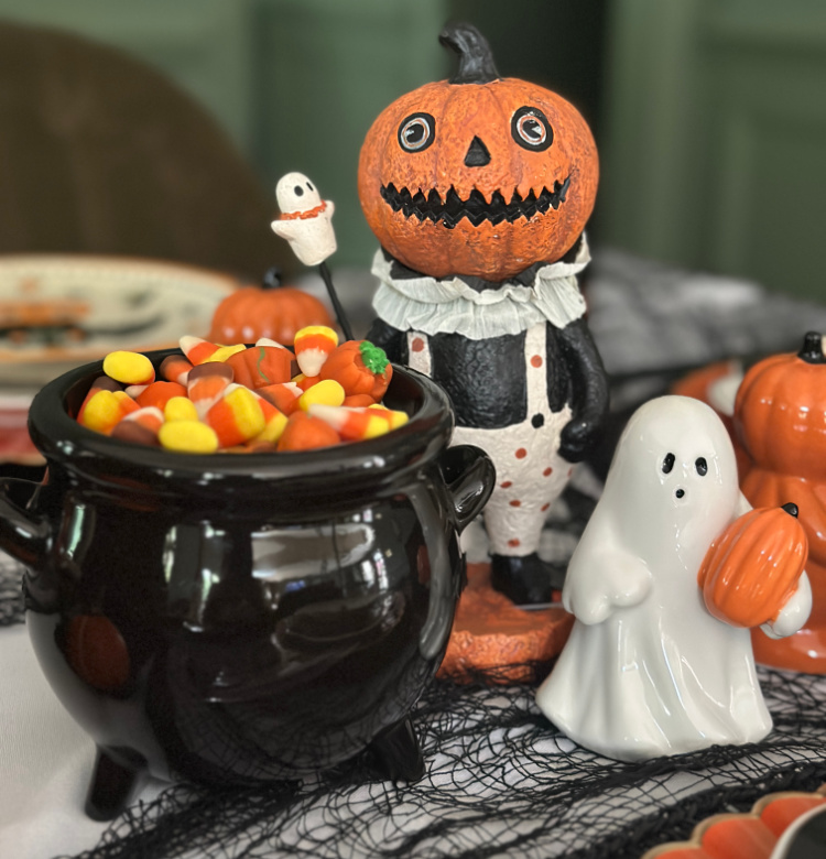 Black cauldron filled with candy corn beside vintage-style jack-o’-lantern and white ghost figurines on a Halloween table.
