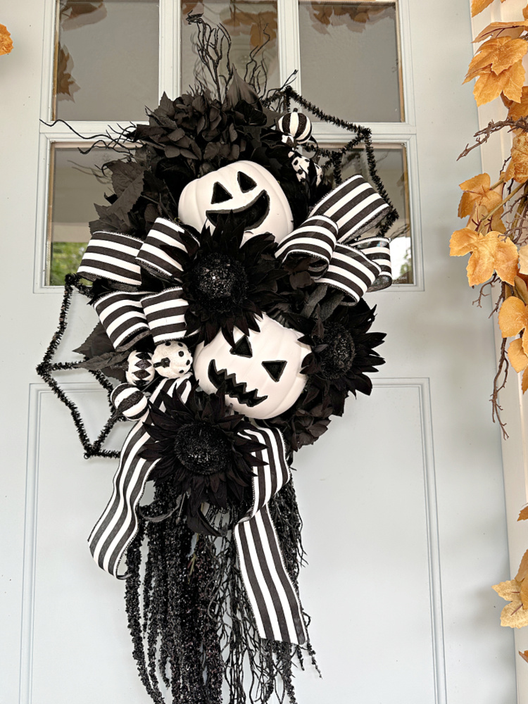 Black-and-white Halloween door swag featuring white jack-o’-lanterns, black flowers and foliage, striped ribbon, spiderweb accents, and trailing black stems.