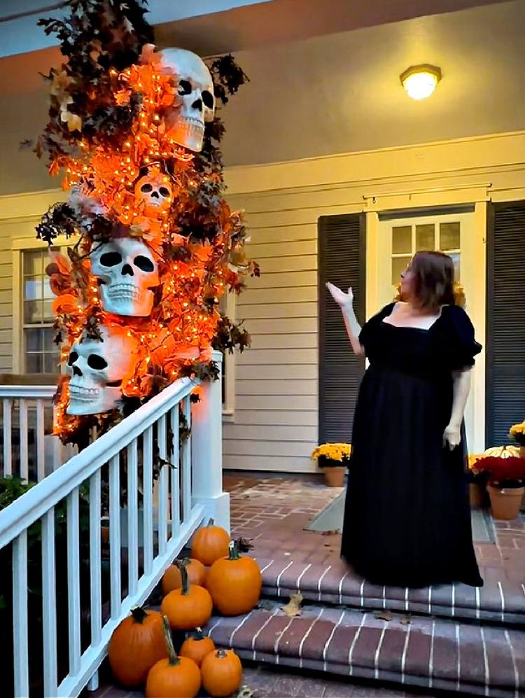 Woman in a long black dress stands beside a towering Halloween porch display filled with oversized skulls, autumn leaves, and glowing orange lights, with pumpkins lining the brick steps.