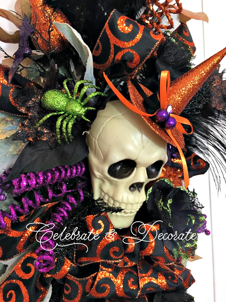 Halloween skull decoration wearing a glittery orange witch hat, surrounded by black and orange ribbon, purple accents, feathers, and a sparkling green spider.