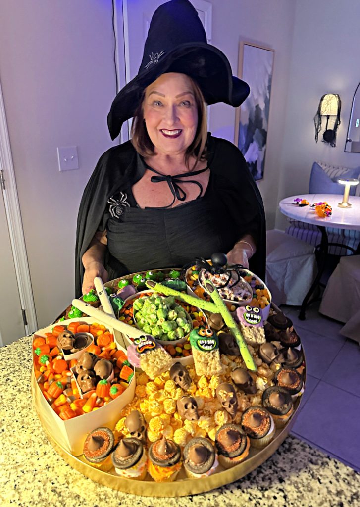 Woman dressed as a witch holds an oversized Halloween grazing board filled with candy, popcorn, cupcakes, chocolate skulls, monster treats, and green dip.