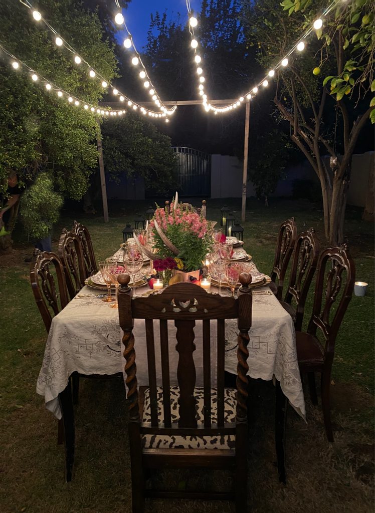 Long outdoor dinner table decorated with flowers and candles beneath glowing string lights in a garden at dusk.