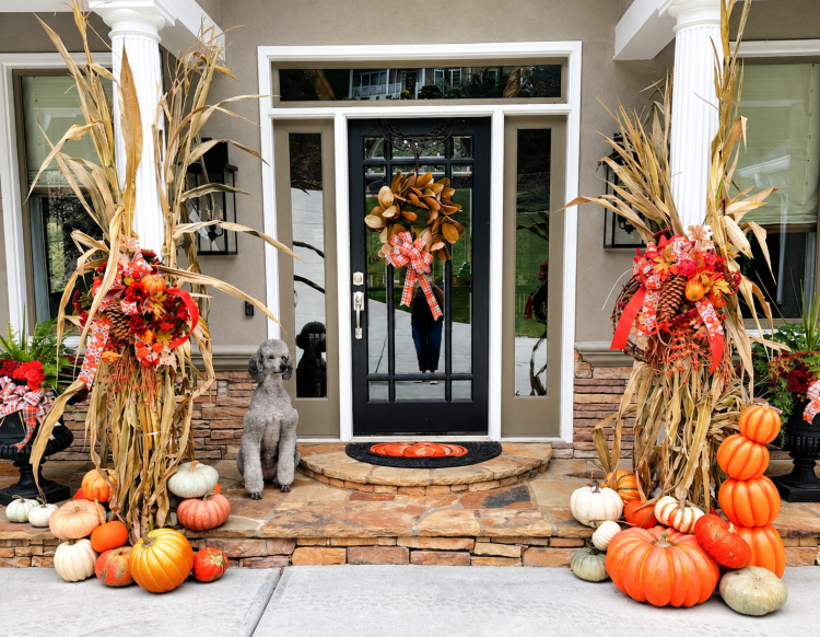 Fall front entry with cornstalks, autumn wreaths and bows, and orange and white pumpkins, with Lily the poodle sitting beside the door.