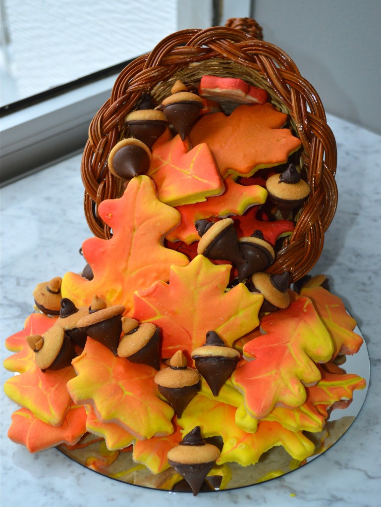 Wicker cornucopia overflowing with autumn leaf cookies decorated in yellow, orange, and red frosting, alongside chocolate and cookie acorns.