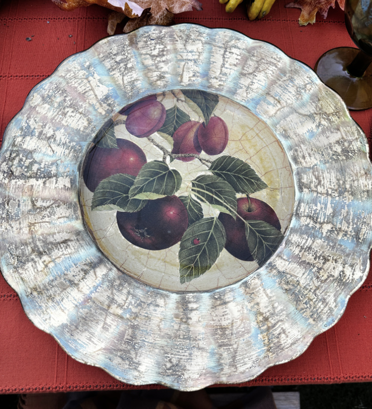 Scalloped charger plate with a distressed metallic rim and a purple plum and green leaf design on a crackled cream center, resting on a rust tablecloth.