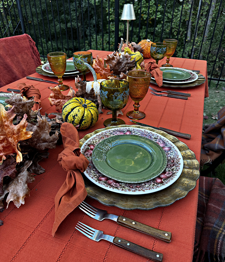 Outdoor fall table with green salad plates layered over fruit-patterned dinner plates and gold chargers, rust knotted napkins, amber and green goblets, and a centerpiece of gourds and autumn leaves.