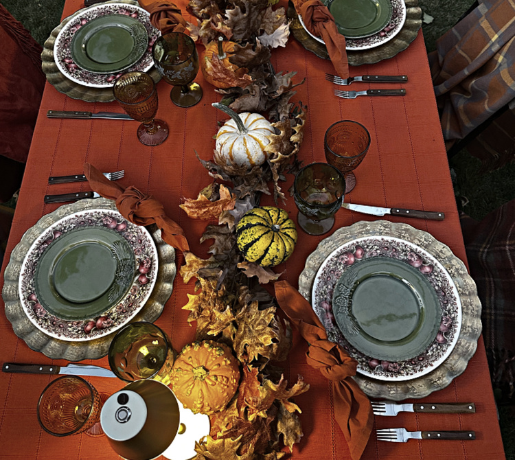 Outdoor fall table with green salad plates layered over fruit-patterned dinner plates and gold chargers, rust knotted napkins, amber and green goblets, and a centerpiece of gourds and autumn leaves.