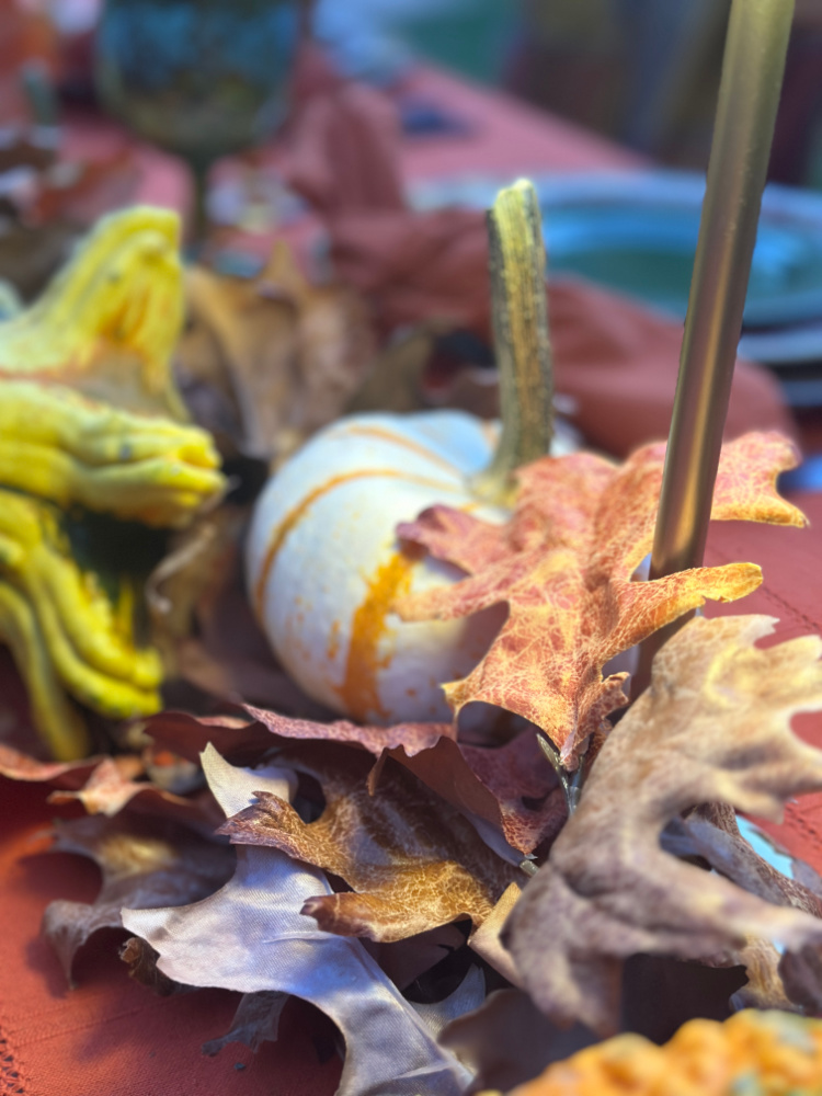Close-up of a white pumpkin with orange stripes nestled among faux autumn oak leaves and yellow-green gourds on a rust-colored tablecloth.