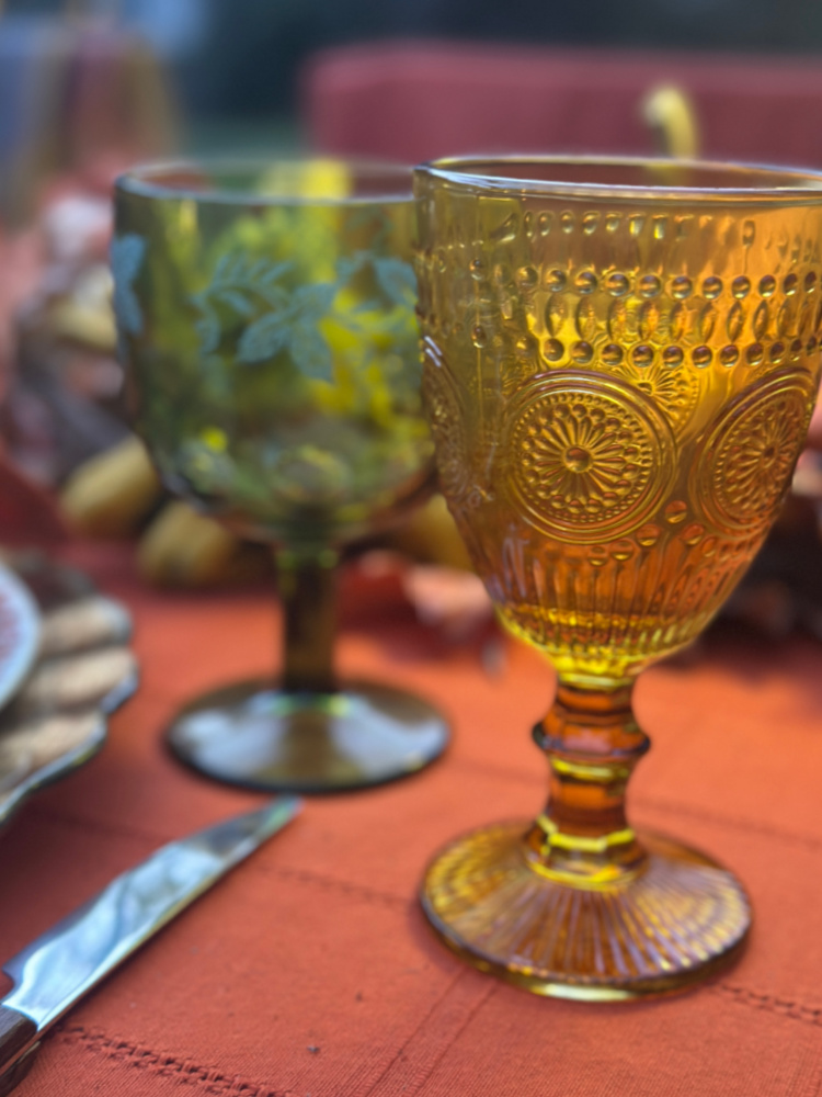 Close-up of an amber goblet with embossed medallion details beside a green botanical-patterned goblet on a rust-colored tablecloth.
