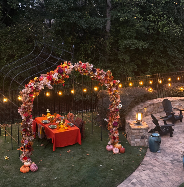 Autumn dining table with an orange tablecloth beneath a metal pergola adorned with fall foliage, flowers, and pumpkins, with glowing string lights and woods behind.