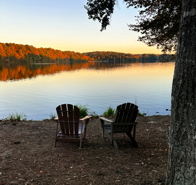Two wooden Adirondack chairs overlooking a calm lake, with golden sunlight illuminating autumn trees and their reflections across the water.