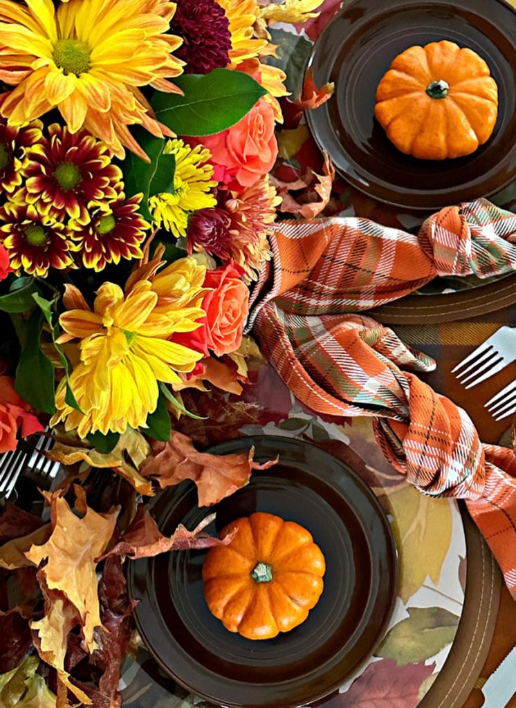 Fall table setting shared on Celebrate and Decorate’s Instagram.  Brown plate with orange mini pumpkin and orange plaid napkin
