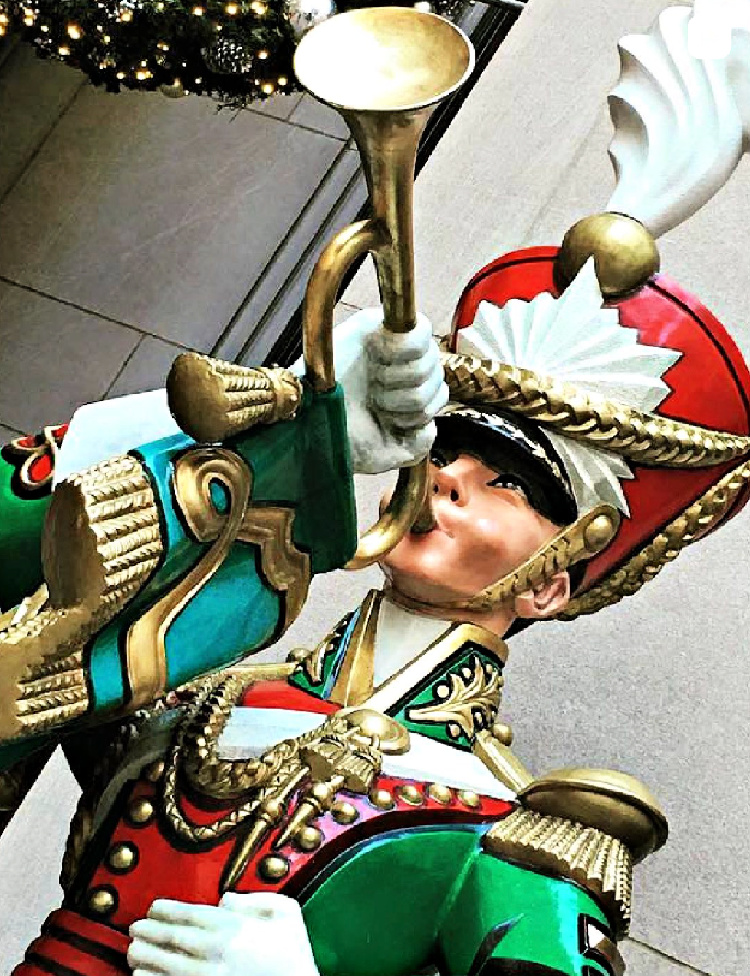 Decorative Christmas toy soldier in a green, red, and gold uniform playing a gold trumpet, with a white-plumed hat and illuminated greenery behind him.