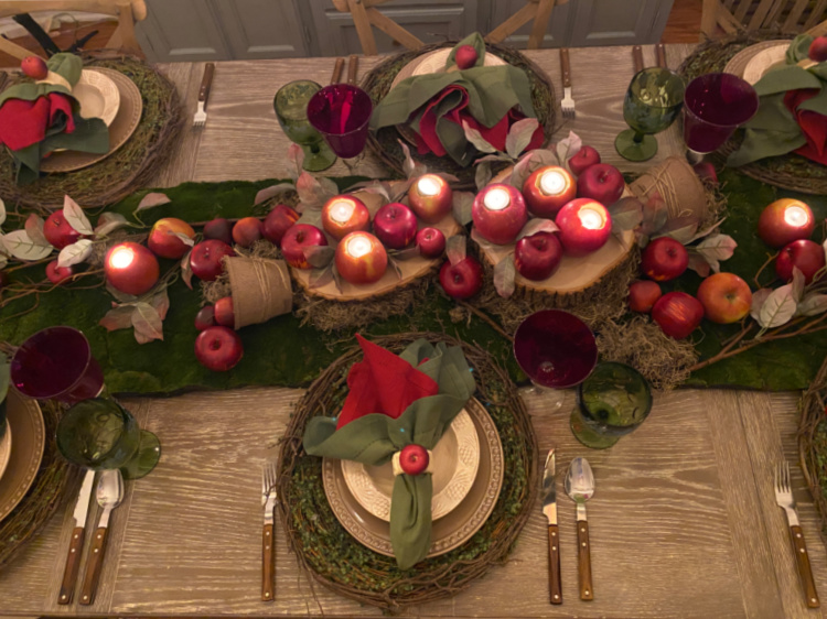 Overhead view of an apple-themed fall table with glowing apple candles, wood slices, and apples on a moss runner, surrounded by twig placemats, layered dishes, red and green napkins, and coordinating goblets.